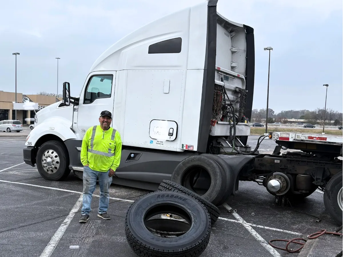 Mobile technician in hi-vis standing beside a semi truck he is servicing on site in a parking lot, with the wheel off and parts laid out