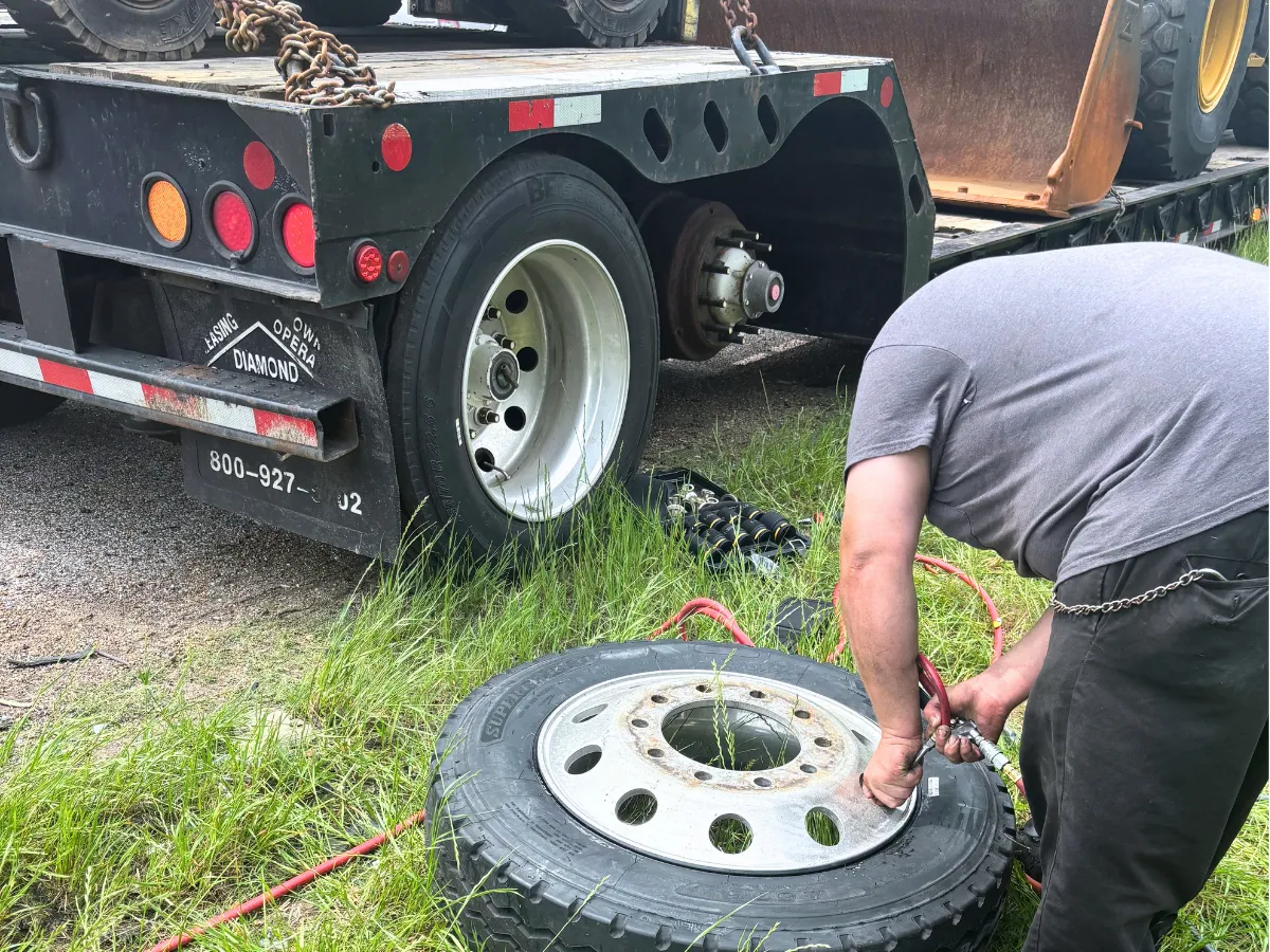 Technician mounting a new trailer tire on the rim up close beside a loaded lowboy trailer